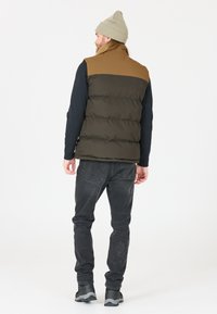 Puffer vest in dark green and brown, featuring a quilted design. Paired with grey jeans and black shoes. Casual beanie on the head.