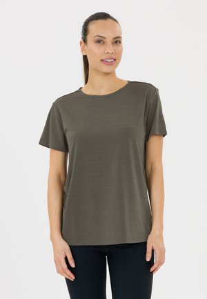 Femme aux cheveux foncés tirés en arrière, vêtue d'un t-shirt à manches courtes vert olive et d'un pantalon foncé, debout devant un fond blanc.