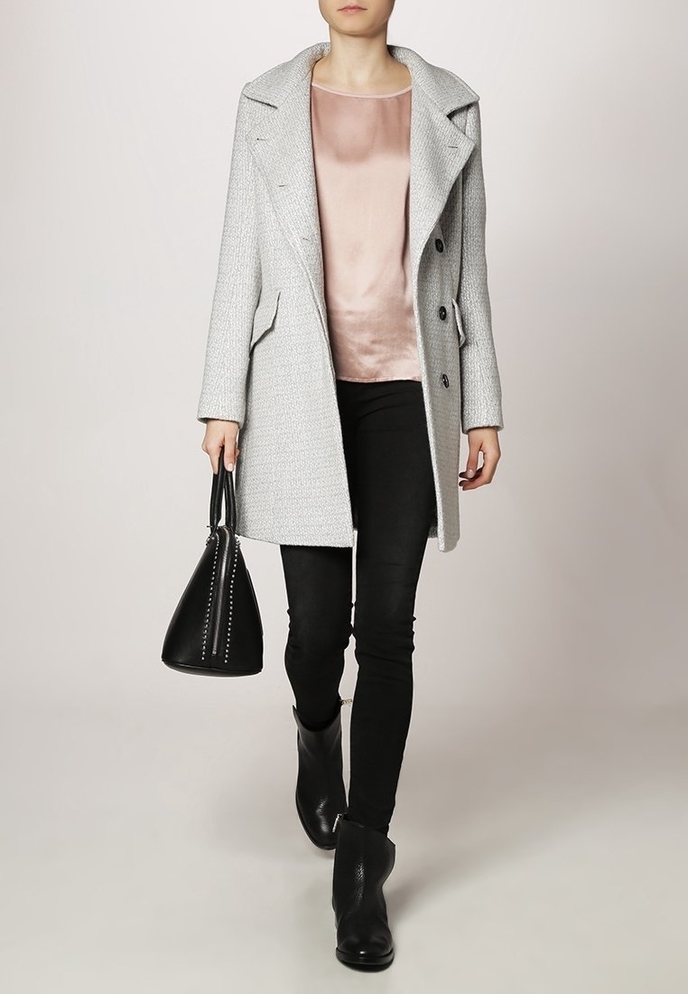 Femme portant un manteau gris clair texturé, une blouse en satin rose, un pantalon noir, des bottines noires, portant un sac à main noir à clous.