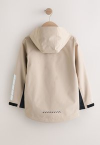 Veste à capuche beige avec doublure intérieure noire et bandes réfléchissantes sur la manche gauche et le bas du dos, suspendue à un crochet en bois contre un mur blanc.