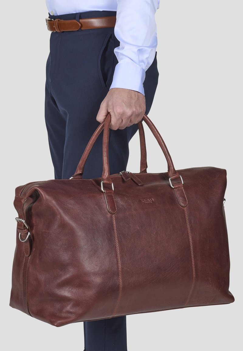 Homme habillé d’un pantalon bleu et d’une chemise blanche tenant un grand sac de voyage en cuir marron par ses poignées.