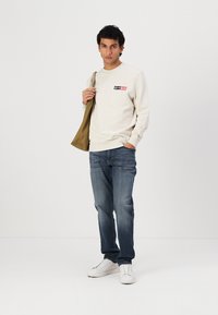 Krem sweatshirt med logo på venstre bryst, blå jeans og hvite sneakers. Modellen holder en brun jakke. Minimalistisk stil, frontperspektiv.