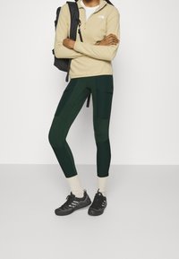 Beige fleece puolipaita, tummanvihreät istuvat legginsit, joissa on tekstuuripaneelit, ja mustat urheilukengät, joissa on harmaita yksityiskohtia.