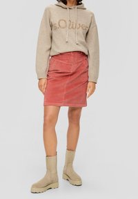Beige huppari, jossa on kultainen script-logo, yhdistettynä punaiseen corduroy-hameeseen. Vaaleat nilkkasaappaat täydentävät asua. Yksinkertainen harmaa tausta.