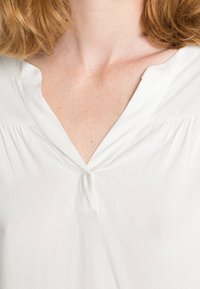 Blusa branca com decote em V, tecido suave, pregas discretas nos ombros e um pequeno detalhe de botão abaixo do decote.