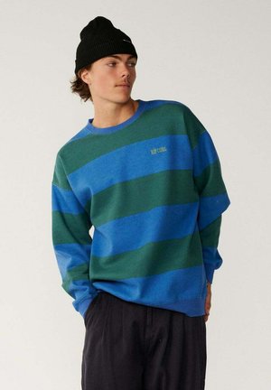 Jeune homme portant un pull rayé bleu et vert et un bonnet noir, debout avec les mains dans les poches, regardant sur le côté sur un fond uni.