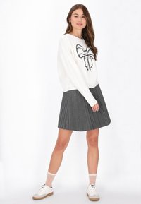 Biały sweter z czarnym haftem w kształcie kokardy, połączony z plisowaną szaro-skalą. Modelka nosi białe trampki i skarpetki w kropki.