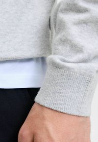 Suéter gris claro con puños acanalados, hecho de una tela suave. Debajo, se visible una capa blanca. El primer plano muestra la textura y los detalles de la costura.