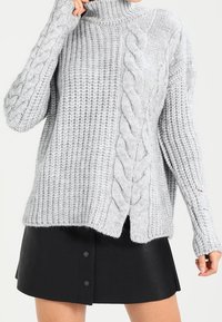 Grau grob gestrickter Pullover mit hohem Kragen, der über Kabelstrickdetails und seitliche Schlitze verfügt, kombiniert mit einem schwarzen Lederrock.