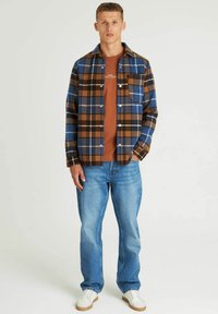 Geruite shirt in blauw, bruin en zwart, met een knoopsluiting aan de voorkant, een borstzak en een textiele stof. Draag het over een bruine t-shirt en blauwe spijkerbroek.