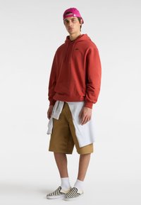 Rote Kapuzenjacke mit einer Fronttasche, graues Crewneck um die Taille gebunden, khakifarbene Shorts und schwarz-weiße Karoschlüpfschuhe. Rosa Kappe.