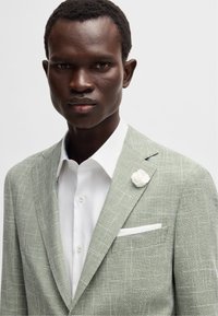 Blazer à carreaux vert clair avec un tissu texturé et un accent de fleur de revers blanche. Porté sur une chemise blanche avec un pochette.