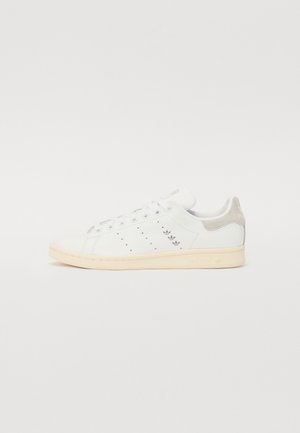 Baskets en cuir blanc avec un onglet de talon en daim beige, un design perforé sur le côté et une semelle en caoutchouc de couleur claire.