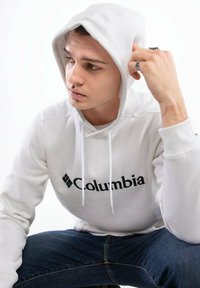 Biała bluza z kapturem z czarnym logo "Columbia", wykonana z mieszanki bawełny, z regulowanymi sznurkami i ściągaczami na mankietach, w połączeniu z niebieskimi dżinsami.
