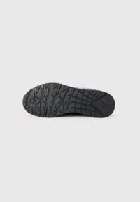 Semelle de chaussure de sport noire avec des motifs de semelle géométriques et en forme de grille sur un fond blanc uni.