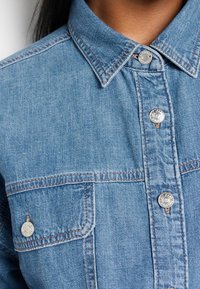 Chemise en jean bleu clair avec col classique, boutons en métal, poches avant et détails de coutures contrastantes. Fini texturé visible.