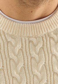 Krämsfärgad kabelstickad tröja med ribbad krage och strukturerat mönster med framträdande vridna stygn. Vit innersidan synlig.