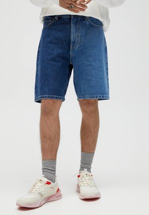 Personne portant un short en denim bleu mi-long, des chaussettes grises, des baskets blanches et rouges, et un haut blanc à manches longues avec les mains jointes au-dessus de la taille.