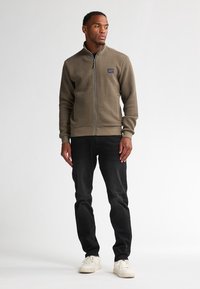 Kaki zip-up jacka med texturerad tyg och ribbade ärmar, i kombination med svarta slim-fit jeans och vita sneakers. Inga synliga mönster eller accenter.