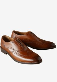 Des chaussures oxford en cuir brun présentent des détails en brogue, une texture lisse et un bout arrondi. Le design comprend un laçage et un talon bas.