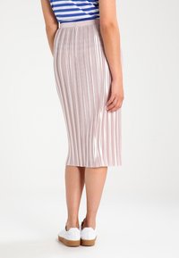 Kvinna iklädd en rosa metallisk plisserad midikjol, en blå och vit randig topp samt vita sneakers, som står mot en vit bakgrund.