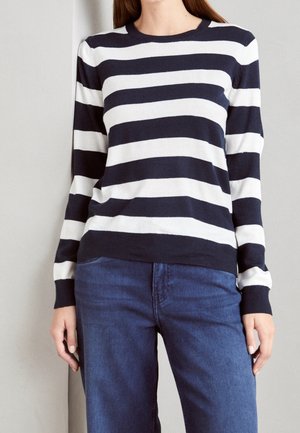 Femme portant un pull à rayures horizontales noires et blanches à manches longues avec un jean bleu, debout contre un mur clair uni.