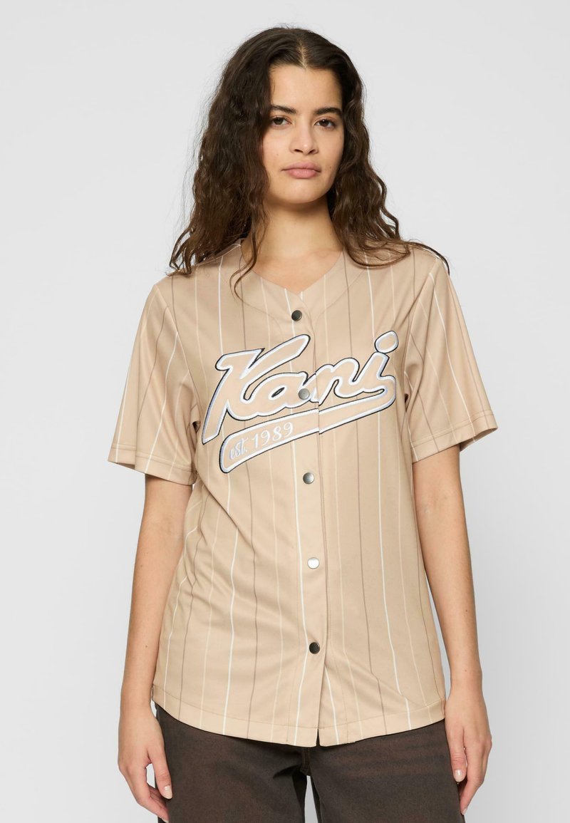 Beige Kurzarm-Jersey mit vertikalen weißen Streifen, Knopfverschluss und einem auffälligen Logo, auf dem "est. 1989" gedruckt ist.