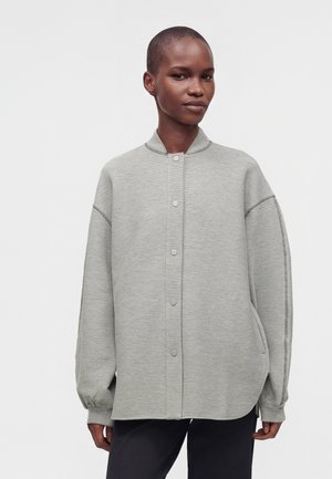 Persona con capelli corti indossa una giacca oversize grigio chiaro con bottoni e maniche lunghe e pantaloni neri su uno sfondo semplice.