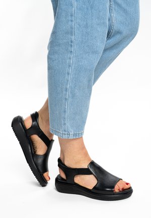 Feet wearing black leather open-toe sandals and light blue cropped jeans against a white background.