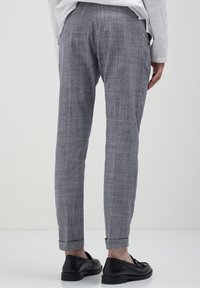 Persona che indossa pantaloni slim-fit grigi a quadri con orli alle caviglie e mocassini in pelle nera, vista da dietro su uno sfondo neutro.