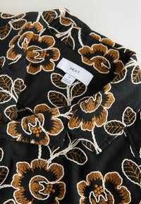 Musta kangas, jossa on ruskea ja valkoinen kirjailtu kukkakuvio sekä vaateetiketti, jossa lukee merkki "NEXT" ja koko "M".