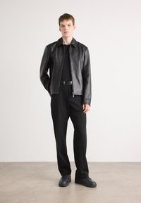 Black leather jacket with a smooth finish, zip closure, and collar, paired with loose black trousers and black shoes. Minimalist design.