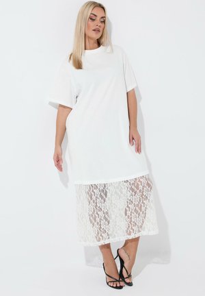 Lyshåret kvinde iført en hvid kjole med korte ærmer og en gennemsigtig blonderkant, kombineret med sorte strop-sandaler med høje hæle.