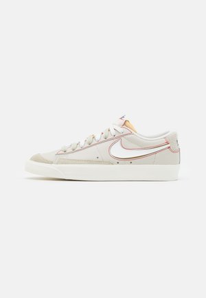 Beiger Sneaker mit weißer Sohle, weißen Schnürsenkeln, roten Nähten und weißem Swoosh-Logo an der Seite.