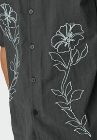 Donkergrijze shirt met korte mouwen, versierd met lichtblauwe bloemenborduursel langs de linkerkant, met vijf zwarte knopen aan de voorkant.