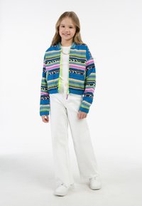 Večbarvna pletena bomber jakna z modrimi, rožnatimi in zelenimi črtami, z zadrgo in rebrastimi manšeti, oblečena z belimi hlačami in supergami.