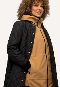 Svart, kviltad jacka med silverdetaljer över en tan färgad zip-up hoodie. Har en hög krage, elastiska muddar och sidofickor.
