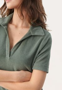 Polo shirt en éponge verte avec une texture douce, manches courtes et col à encoches. Gros plan sur le cou et les détails des épaules.