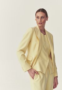 Femme aux cheveux plaqués en arrière, portant un costume sur mesure jaune pâle composé d'un blazer, d'un gilet boutonné et d'un pantalon assorti, regardant vers la gauche.