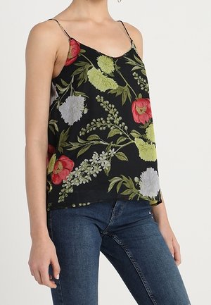 Kobieta w czarnej, bezrękawnikowej bluzce w kwiaty w kolorach czerwonym, żółtym i białym, połączonej z niebieskimi dżinsami na jednolitym tle.