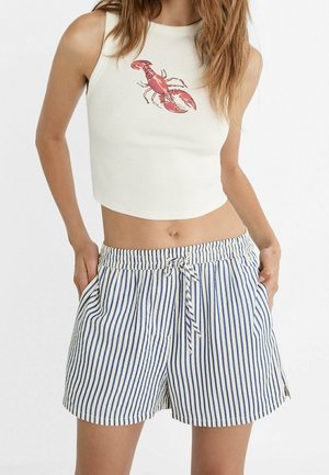 Femme portant un crop top blanc avec un motif homard rouge et un short à rayures verticales bleu et blanc avec une taille à cordon.