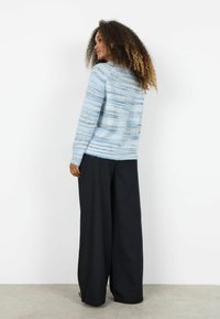 Femeie cu păr creț, purtând un pulover albastru cu dungi și pantaloni largi negri, stând în picioare și privind către un perete alb.