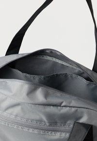 Šedá plátěná taška se černými popruhy, s horním zipem a vnitřní kapsou. Hladká textura a pevná konstrukce.