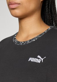 Nærbillede af en person iført en sort Puma t-shirt med leopardmønstret pynt omkring halsudskæringen og Puma-logoet broderet på brystet.