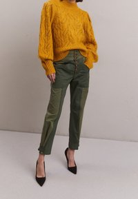 Mosterdkleurige gebreide trui met patroon, hoogwaist olijfgroene broek met voorste knopen en zwarte puntige hakken.