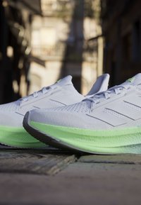 Gros plan de chaussures de course blanches et vert clair posées sur une rue pavée avec des bâtiments flous en arrière-plan.