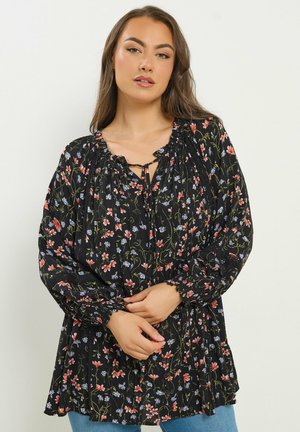 Donna con capelli lunghi castani che indossa una camicetta nera a fiori rossi e blu, in piedi davanti a uno sfondo chiaro e uniforme.