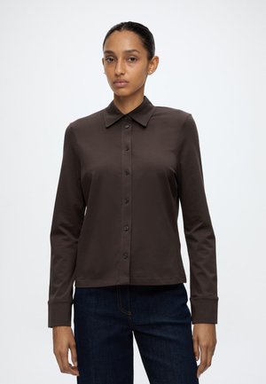 Femme aux cheveux foncés tirés en arrière porte une chemise marron foncé à manches longues boutonnée et un jean bleu foncé sur un fond clair uni.