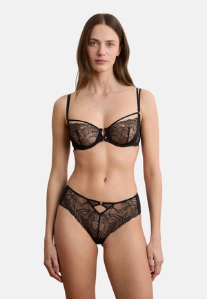 Vrouw draagt zwarte kanten beha en bijpassende hoge taille kanten slipjes, staand tegen een effen lichte achtergrond, met het gezicht naar voren gericht.