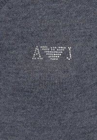 Grå stickad tyg med en texturerad logotyp av små, glänsande nitar. Designen är enkel och minimalistisk och visar en jämn konsistens.